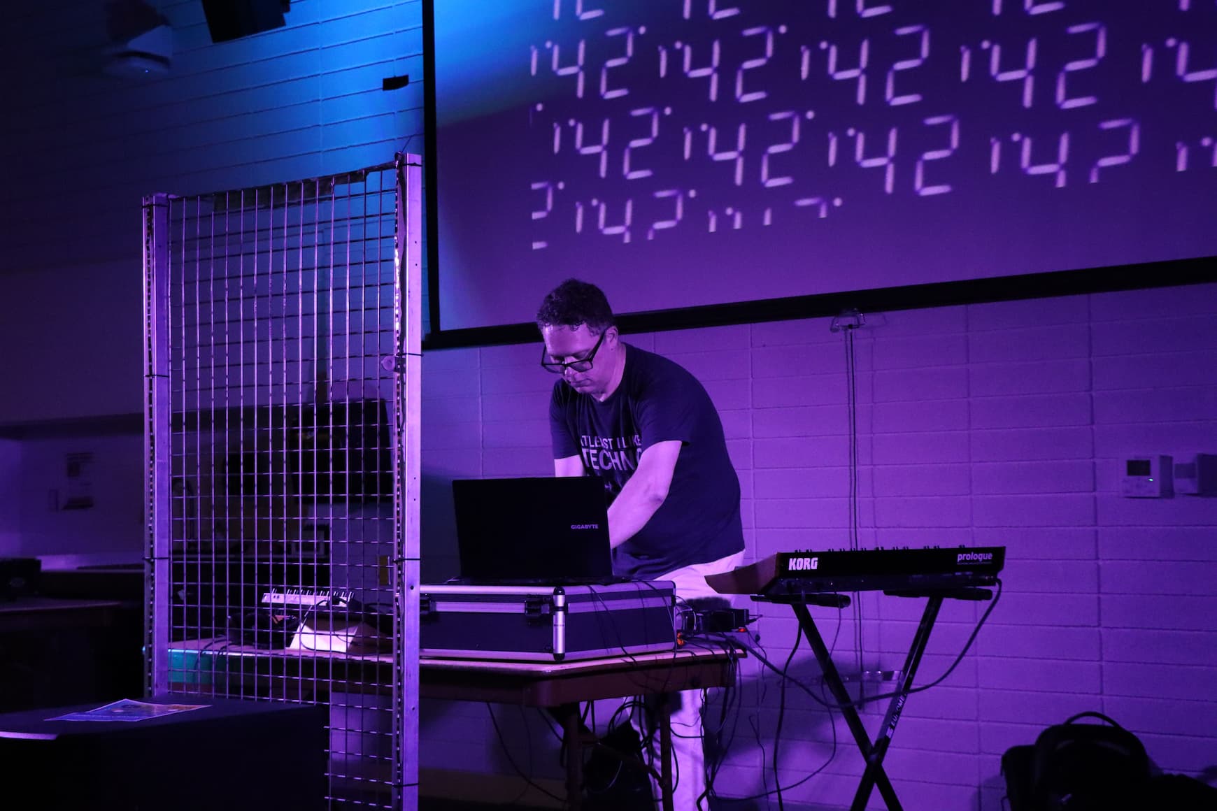 artist performing live with a laptop and Korg synthesizer under purple stage lighting, numbers projected on screen behind them
